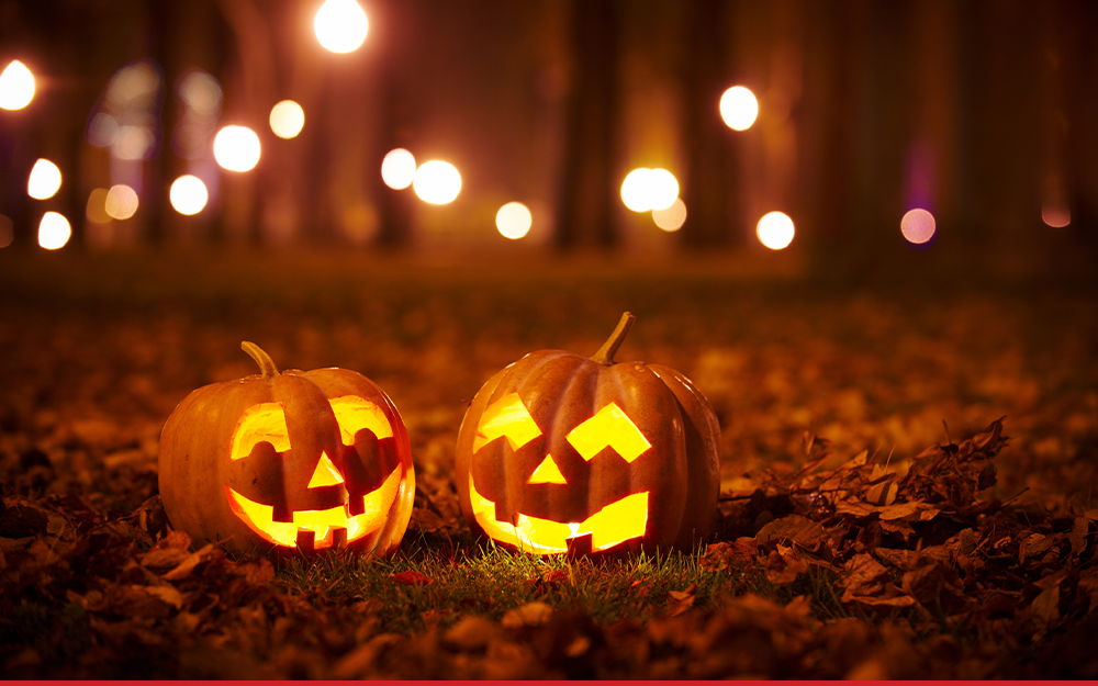 Two pumpkins carved with scary faces sit on the forest floor. Behind them, there are bokeh lights between trees.