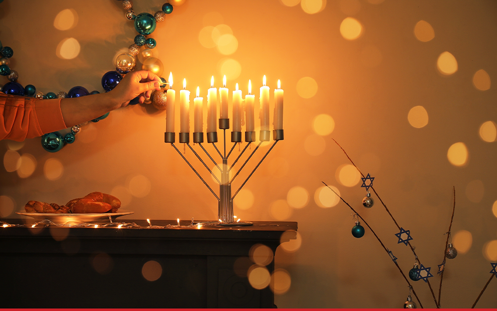 An arm reaches in to frame to light a menorah. There is a mirror hanging on the wall and lots of bokeh lights around with menorah.