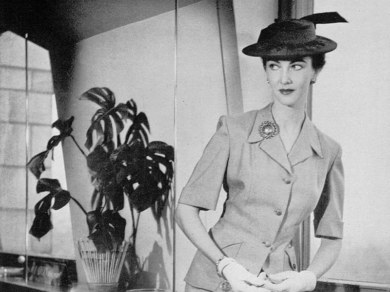 Black and white photograph of a woman stood next to a plant.