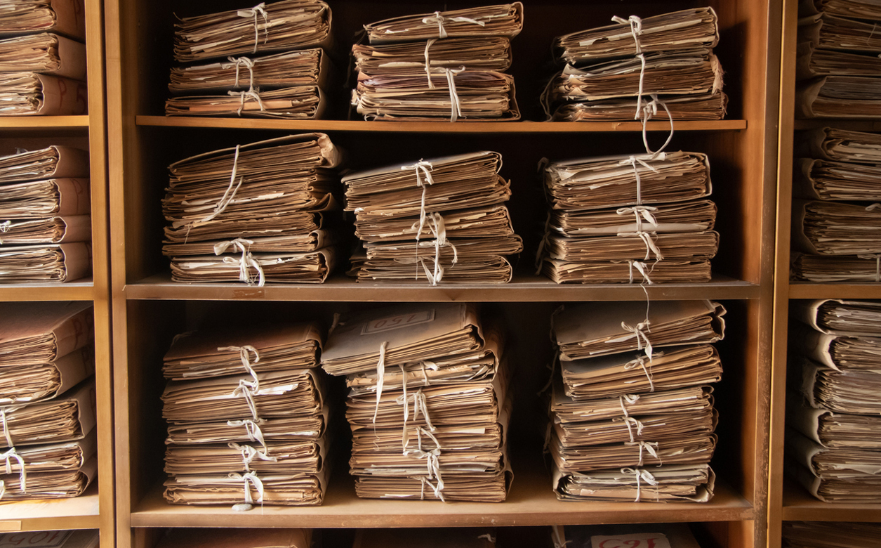 A shelving unit with piles of archival documents stacked neatly, tied with a white ribbon and placed in rows on the shelves.