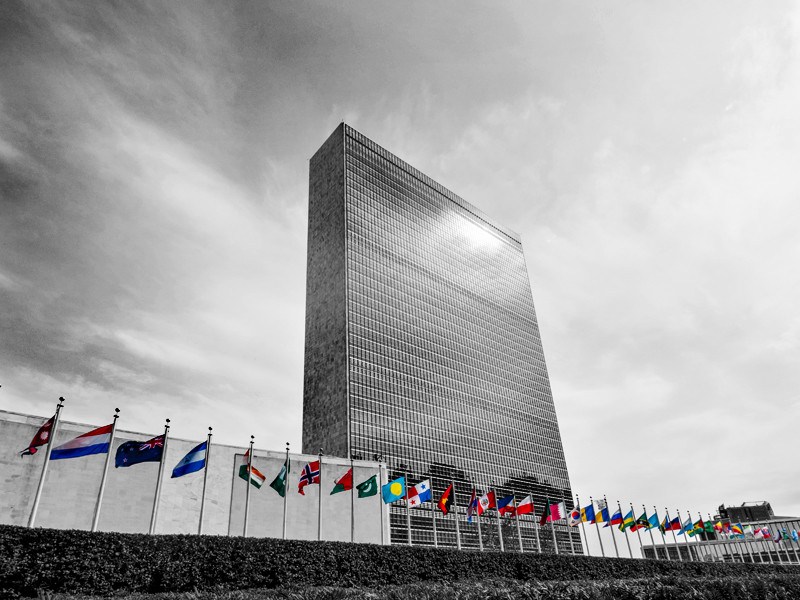 A black and white image of the United Nations Secretariat Building with flags from different nations in colour in front of the building.