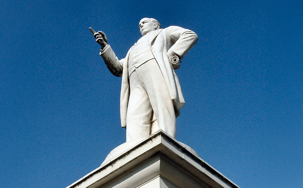 Charles Bradlaugh Statue Northampton, the status is made of white stone and is set against a blue sky from an upward angle.