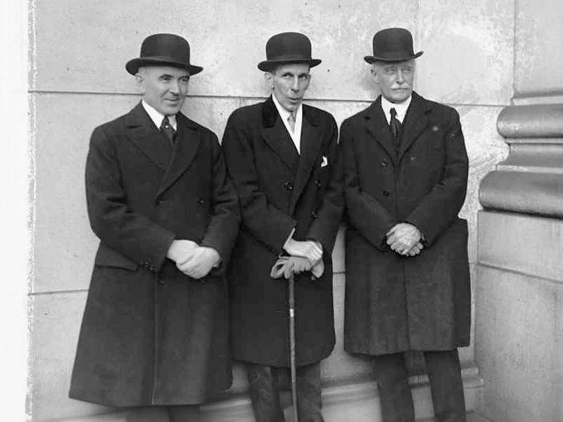 Smiddy, Massey, and Howard, in hats, stood against a stone wall