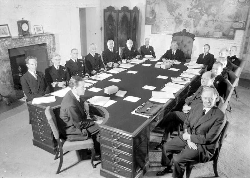 The Board of Admiralty meets in a board room. They are all seated around a table, looking at the camera.