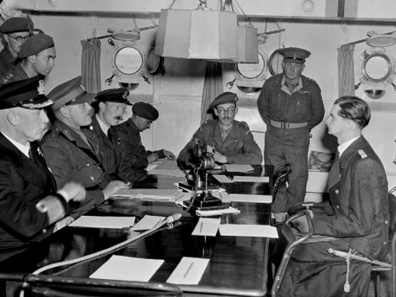A scene on board HMS BULLDOG during the first conference with Captain Lieutenant Zimmerman prior to the signing of the surrender document which liberated the Channel Islands. Nine men gather around a table.