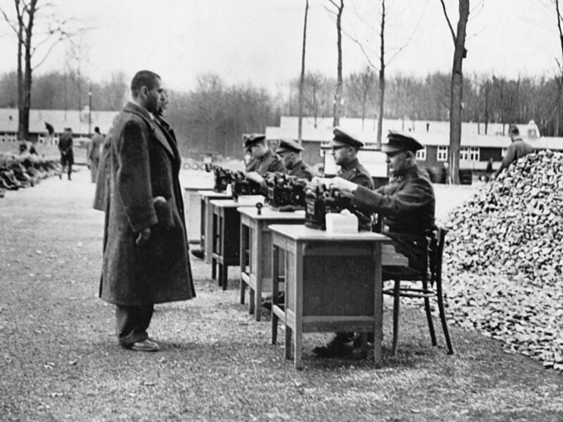 Gestapo officials recording data on incoming prisoners at a German concentration camp. Many others are seated on the ground.