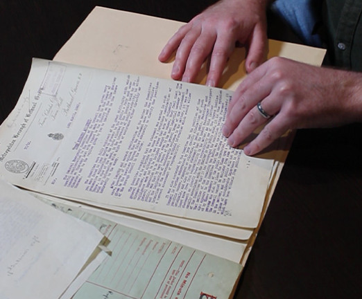 A photo of Dr Charlie Hall leafing through several documents from a file at the National Archives. The documents relate to the Bethnal Green Tube Disaster.