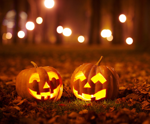 Two pumpkins carved with scary faces sit on the forest floor. Behind them, there are bokeh lights between trees.