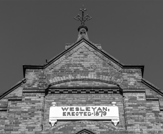A black and white photo of the top of a Church. A sign on the Church reads Wesleyan Erected 1879.