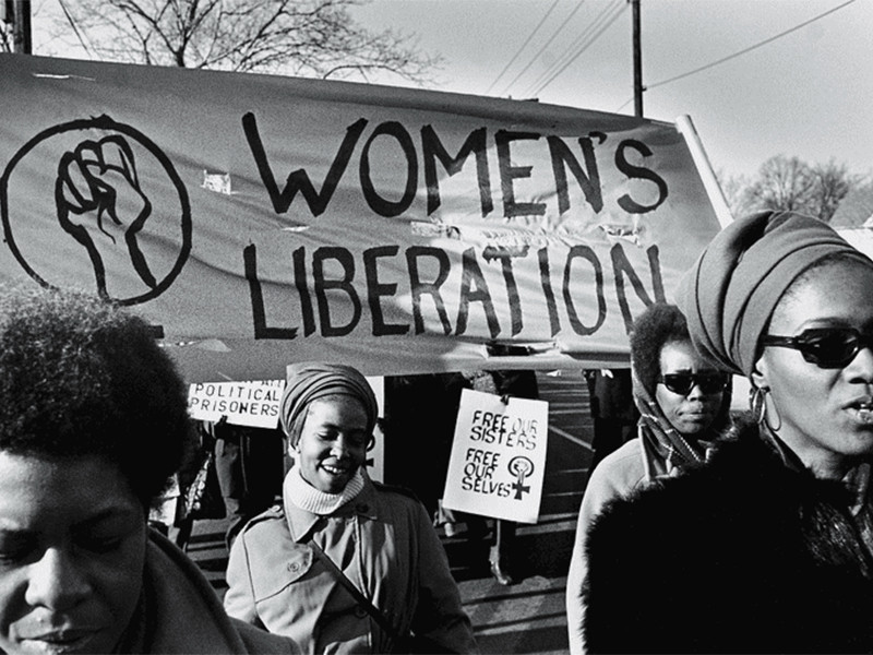 Black and white photograph of women marching, featuring a prominent banner with the text Women's Liberation and a drawing of a closed fist.