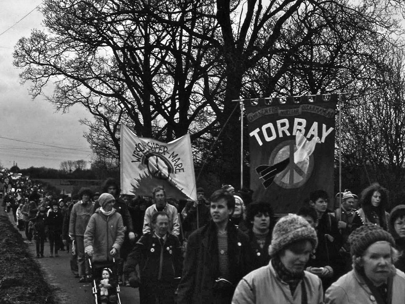 Black and white photograph of women marching, featuring two prominent banners with peace signs on them. One says Campaign for Nuclear Disarmament Torbay, the other says Weston-super-Mare.