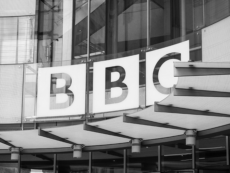 A black and white image of a BBC building, zoomed in on the BBC logo, which is on curved windows.