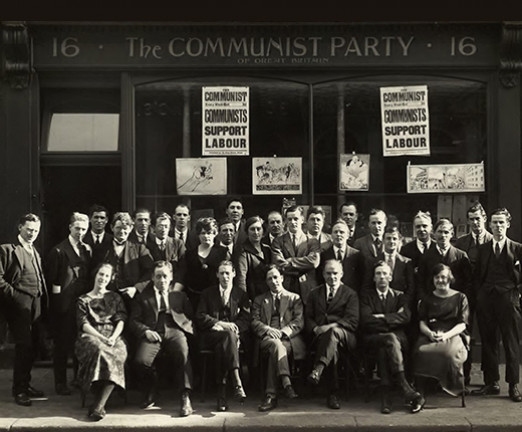 A black and white photo of a group of people sat in front of a building. On the building it says The Communist Party.