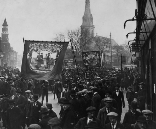 A black and white photo of a busy street with lots of people walking through it. There is a cloth banner being held up.