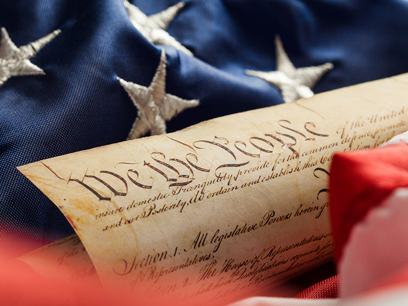 An image of the US constitution laid on top of a USA flag. On top of the constitution in big letters it says 'We the People'.