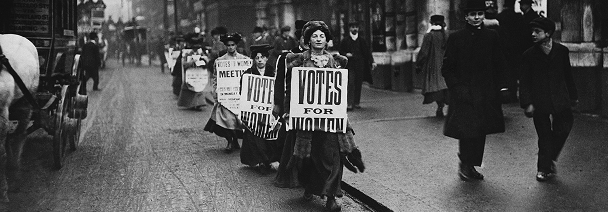 A line of women with sandwich boards advertising a suffragettes meeting in 1912. The woman at the front holds a sign reading 'Votes for Women'.