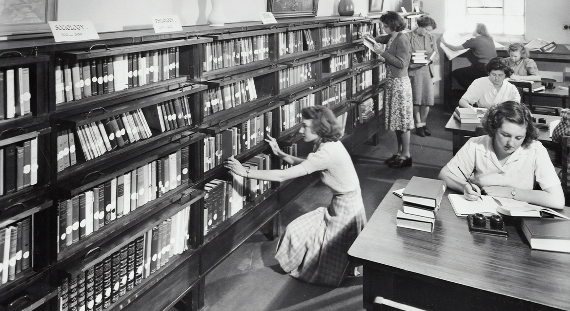 A black and white image of a library with women choosing books and sat at tables reading and writing.