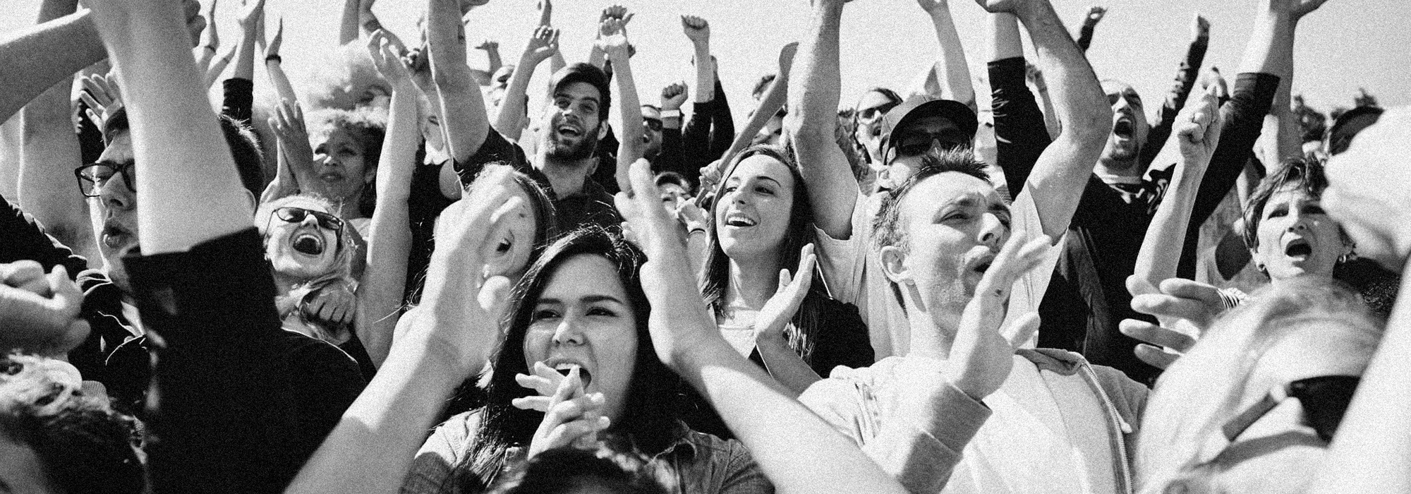 A black and white image of a crowd of people cheering and clapping their hands above their heads.