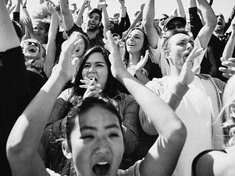 A black and white image of a crowd of people cheering and clapping their hands above their heads.