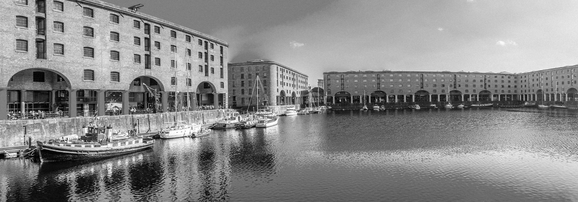 A black and white image of Liverpool docks. The docks are surrounded by buildings and small boats are parked round the edges of the docks.