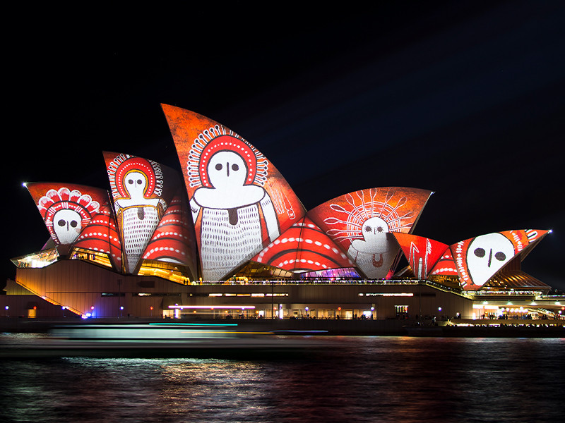 A photograph of Sydney Opera House illuminated in lights for 'Vivid Sydney' a light festival with abstract designs, vivid colours and aboriginal artwork.