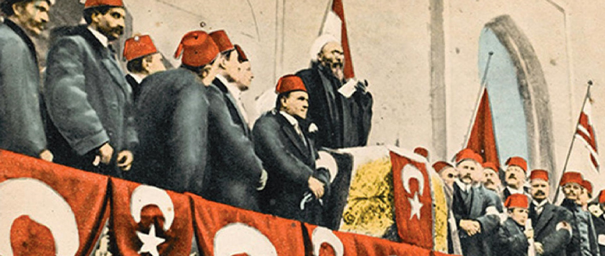 An image of the Ottoman declaration of Jihad, 1914. People are gathered on a balcony wearing hats with the Ottoman flag on. In the middle is a podium where someone is speaking whilst holding a piece of paper.