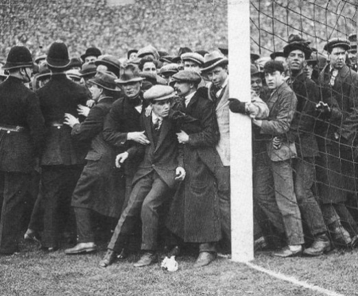 A large crowd of people stood next to a football pitch. Police officers to the left are trying to control the crowd.
