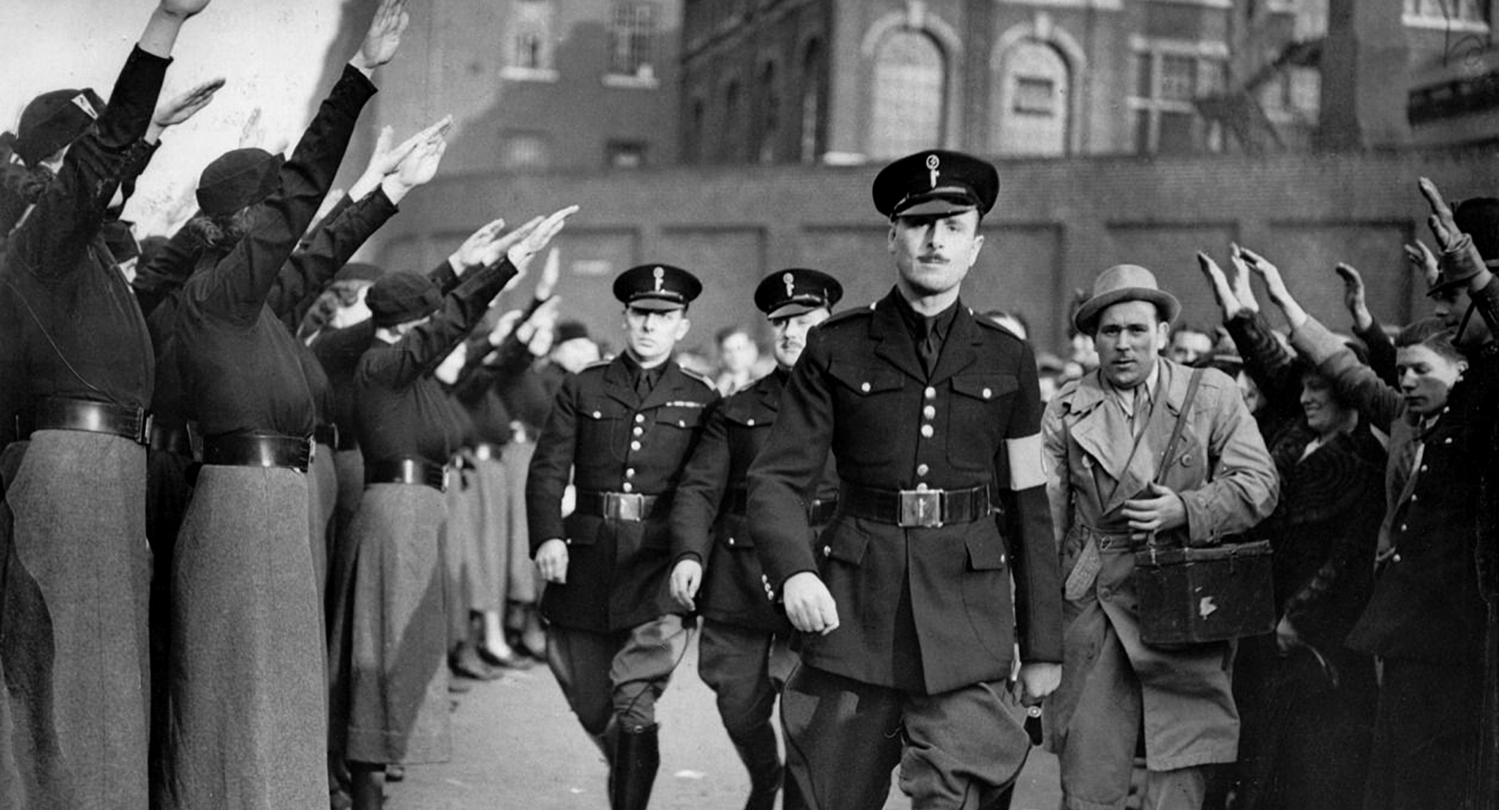 An image of Oswald Mosley walking towards the camera. To the left of him are a line of people saluting to him.