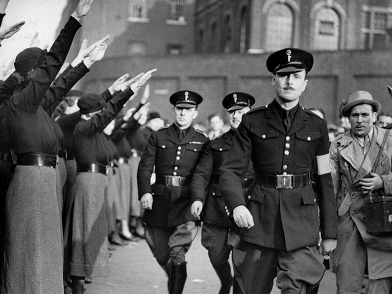 An image of Oswald Mosley walking towards the camera. To the left of him are a line of people saluting to him.