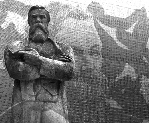 A black and white photograph of the Friedrich Engels statue in Manchester. A mural of his face is also on the wall behind the statue.
