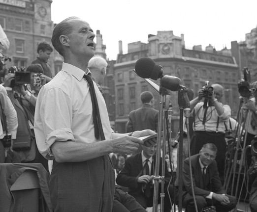 A black and white photograph of John Gollan giving a speech into microphones.