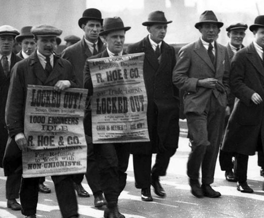 A black and white photograph of workers on strike, holding up posters and marching.