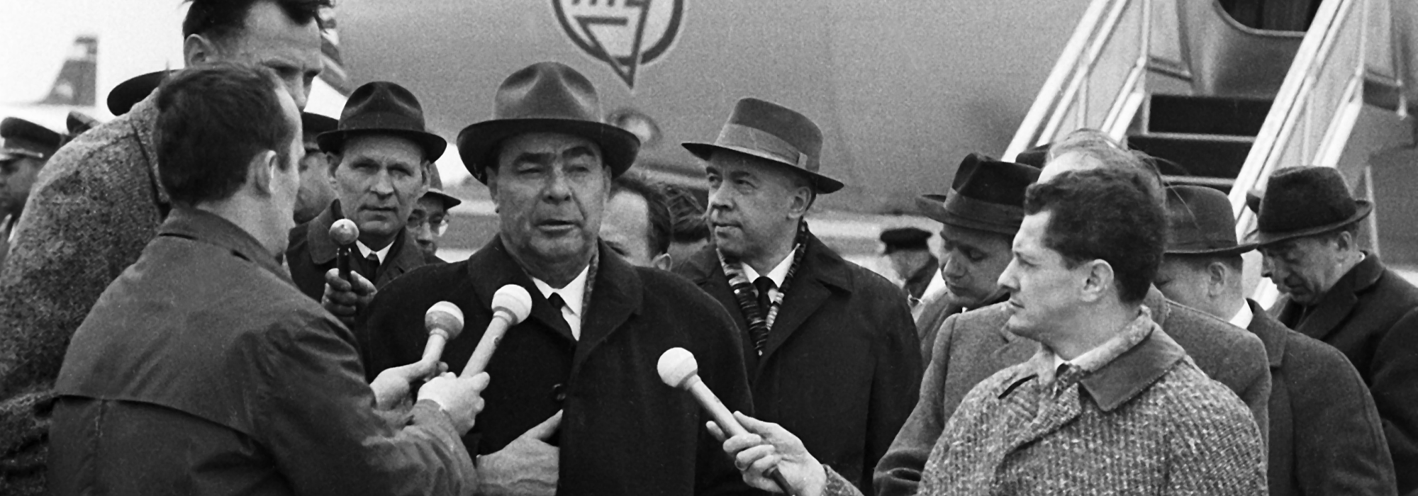 A black and white image of Leonid Ilyich Brezhnev, General Secretary of the Communist Party of the Soviet Union, speaking into microphones which people are holding up to his face.