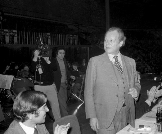 A photograph taken at the signing of the Basic Treaty in Berlin. A man stands at a table while those seated clap. A video camera films him, and an orchestra is behind them.