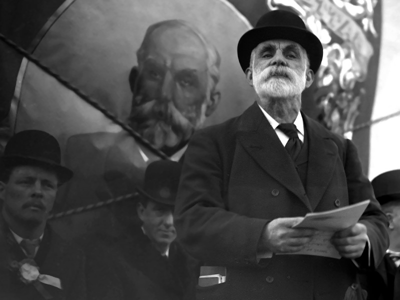 A black and white image of a man with a beard, wearing a hat. He is holding paper in his hands.
