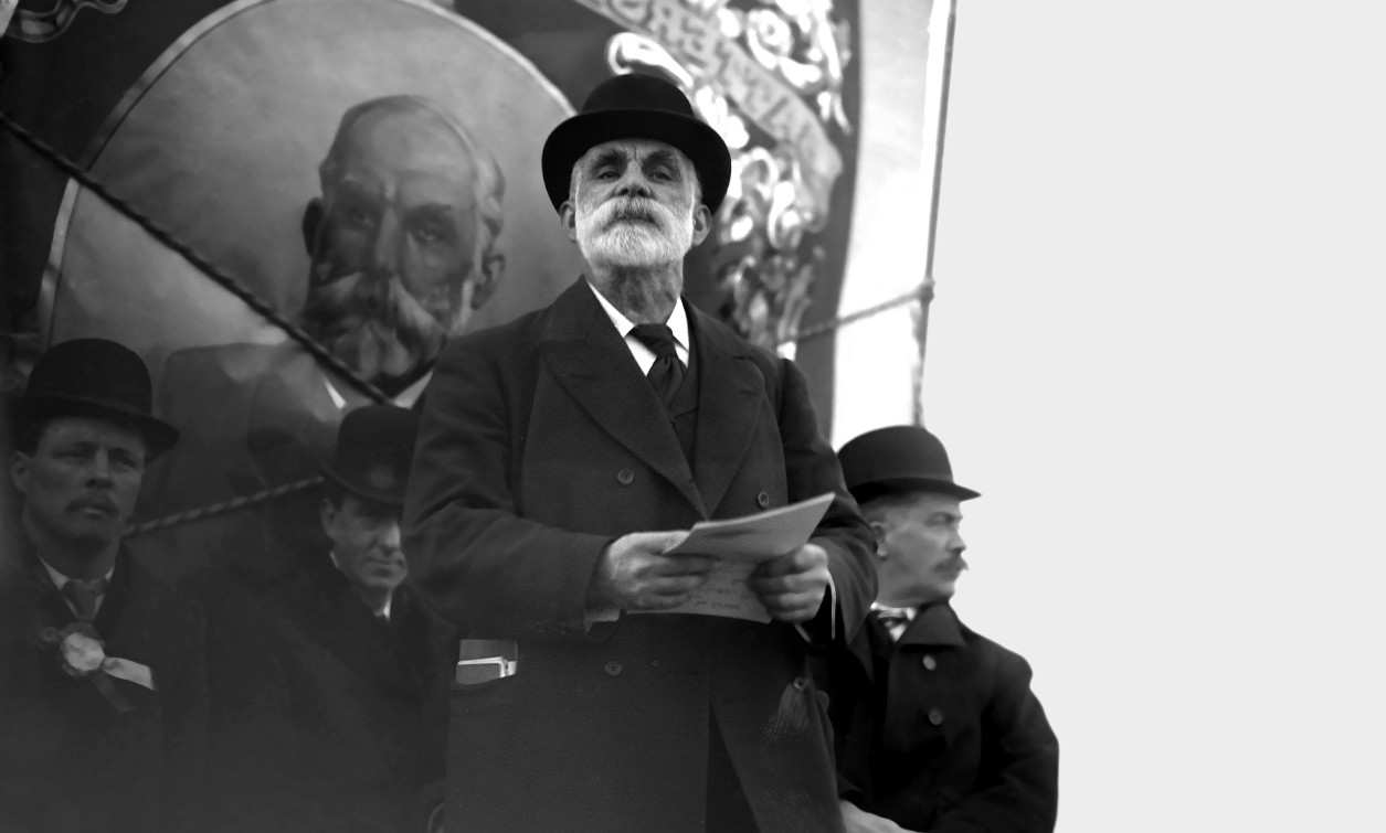 A black and white image of a man with a beard, wearing a hat. He is holding paper in his hands.