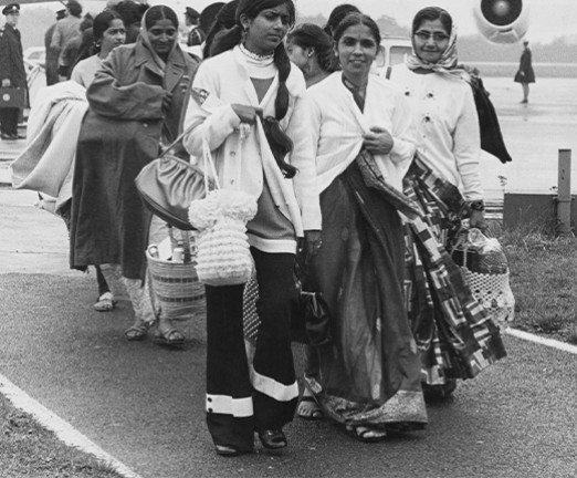 A photograph of several Asian women airlifted from Uganda on a charter flight after their arrival at Stansted Airport, 1972. They walk away from the plane.