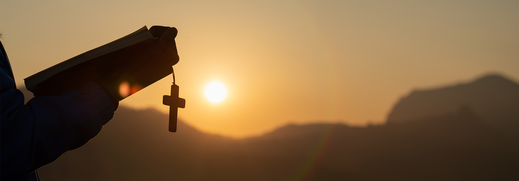 Person holding a holy bible and cross in their hands and praying as the sun rises.