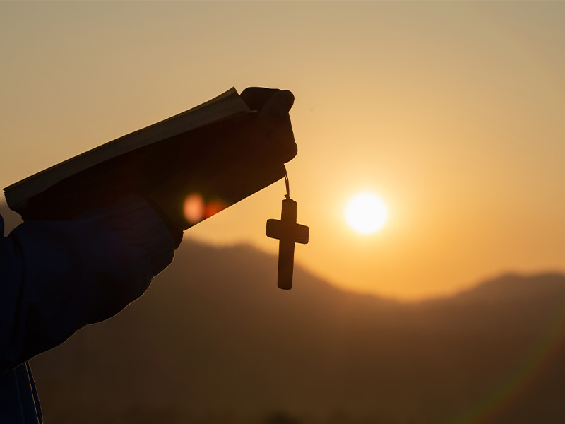 Person holding a holy bible and cross in their hands and praying as the sun rises.