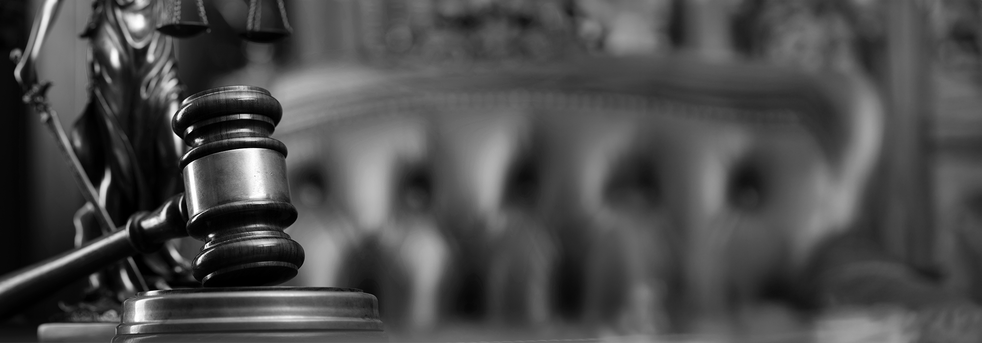 A black and white image of a gavel resting on a table, with a plush chair and scales of justice in the background.