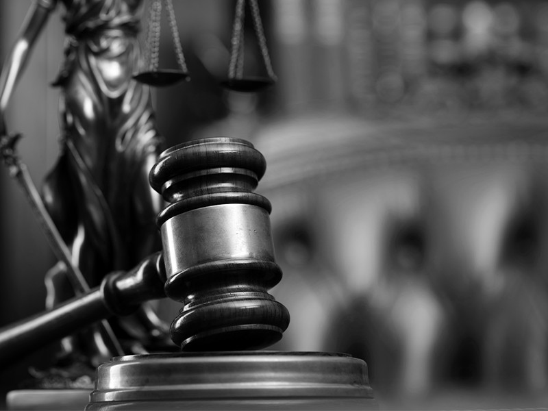 A black and white image of a gavel resting on a table, with a plush chair and scales of justice in the background.