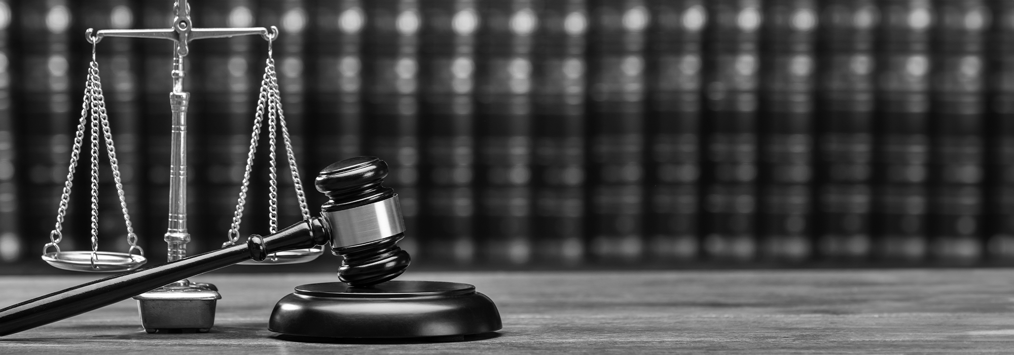 A black and white image of a gavel resting on a table, with a row of books and scales of justice in the background.