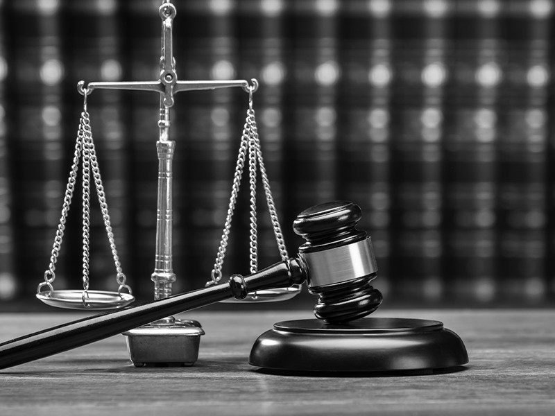 A black and white image of a gavel resting on a table, with a row of books and scales of justice in the background.