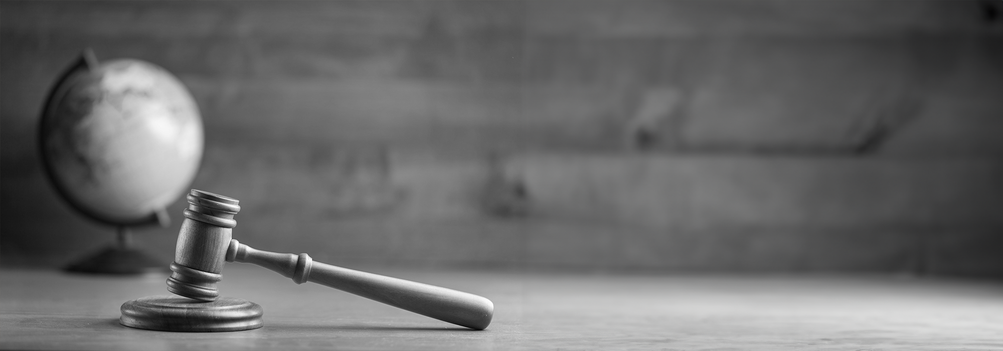 A black and white image of a gavel resting on a table with a globe in the background.