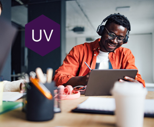 A photograph of a man smiling while writing on an electronic tablet. He sits at a table covered in stationary, and wears headphones. A purple hexagon featuring the letters UV appears on the left of the image.
