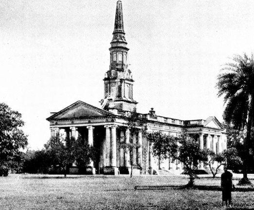 A black and white photo of a church with a large spire nestled within trees. A person in the foreground looks towards the building.