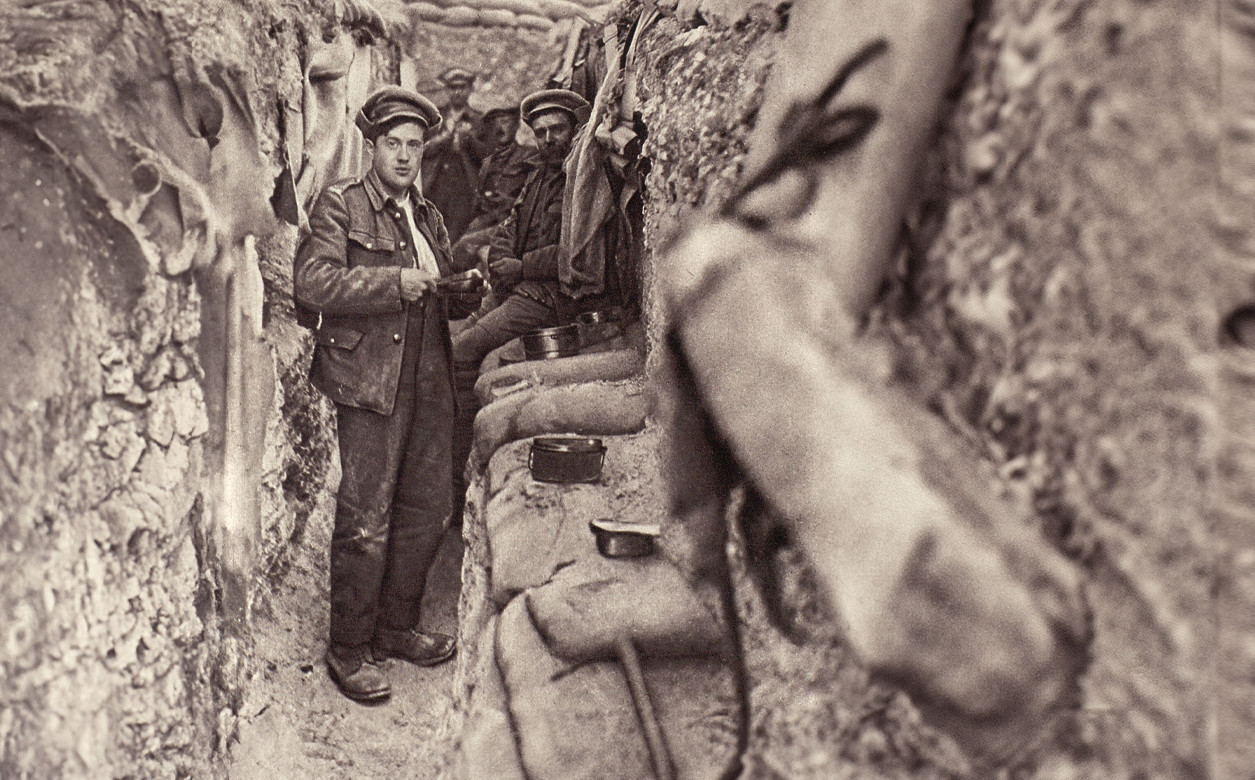An image taken from within a trench. A soldier stands and looks directly at the camera, with other soldiers behind him.