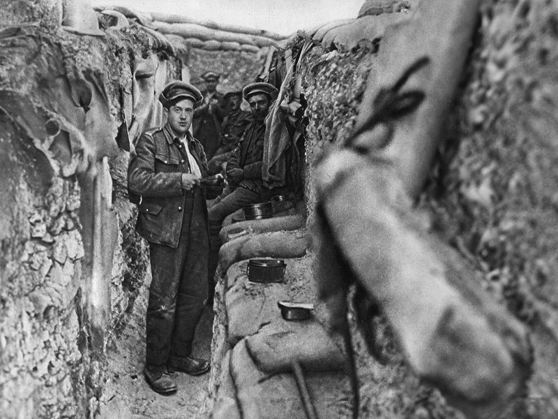 An image taken from within a trench. A soldier stands and looks directly at the camera, with other soldiers behind him.