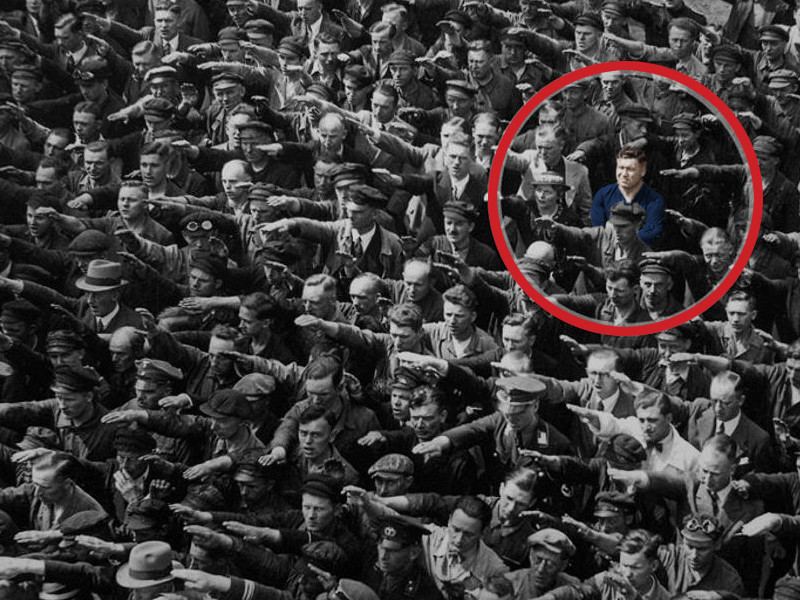 An image of a crowd of people all performing the Nazi salute. Highlighted in a red circle is a man refusing to salute, showing his defiance and support of anti-fascism.