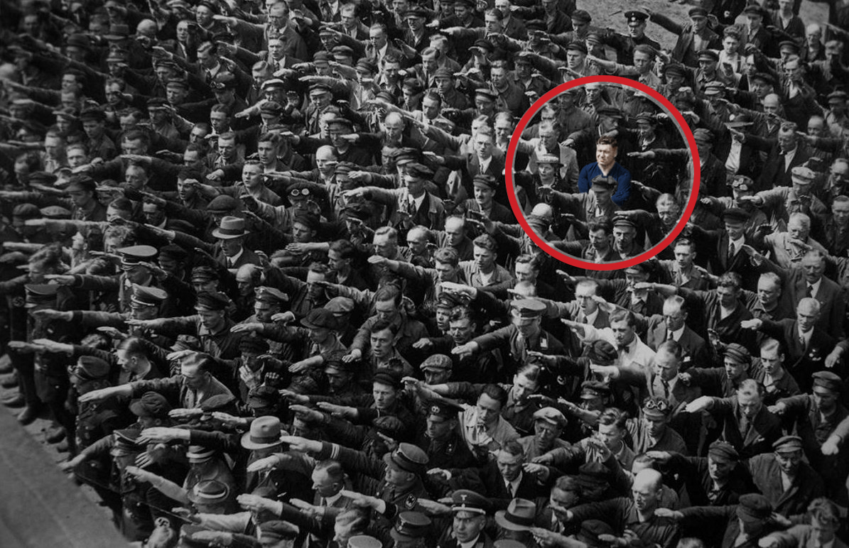 An image of a crowd of people all performing the Nazi salute. Highlighted in a red circle is a man refusing to salute, showing his defiance and support of anti-fascism.